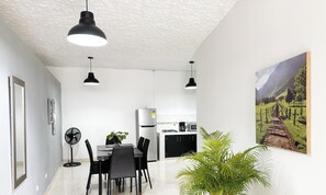 Une cuisine moderne avec des meubles blancs, un rĂ©frigĂ©rateur et une machine Ă cafĂ©. On y trouve un coin repas avec une table et des chaises, ainsi quâun ventilateur au sol.