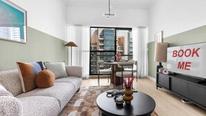 A modern living room with a sofa, a coffee table, a TV, and a view of the city through large windows. - Stayshort - Business Bay for 2 With Water Views (Dubai)