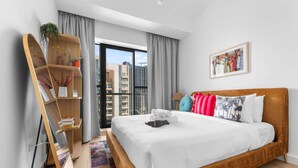 A modern hotel room with a large bed, a wooden side table, a bookshelf, and a city view. - Stayshort - Business Bay for 2 With Water Views (Dubai)