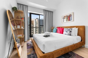 A modern hotel room with a large bed, a wooden side table, a bookshelf, and a city view. - Stayshort - Business Bay for 2 With Water Views (Dubai)