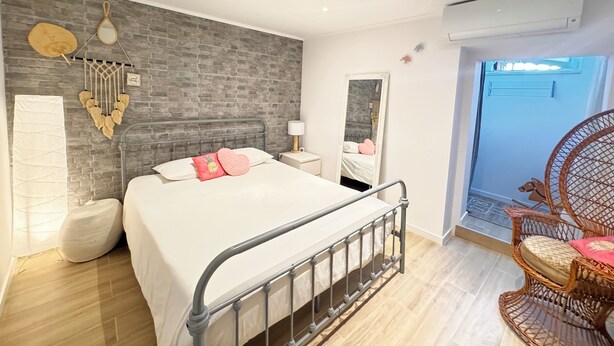 Ein modernes Schlafzimmer mit einer Wand im Ziegelstein-Design, einem Bett mit weißer Bettwäsche und rosafarbenen Kissen, einem Nachttisch mit Lampe, einem großen Spiegel und einem Korbstuhl.