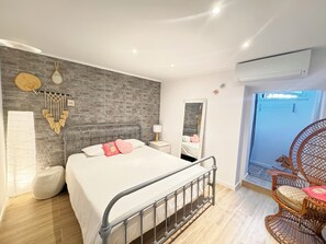 Un dormitorio moderno con pared de ladrillo, una cama con ropa blanca y almohadas rosas, una mesita de noche con lámpara, un espejo grande y una silla de mimbre.
