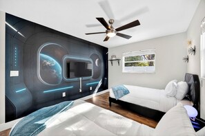 A modern bedroom with a large wall mural, a ceiling fan, a window with a view, and a bedside table with a lamp.