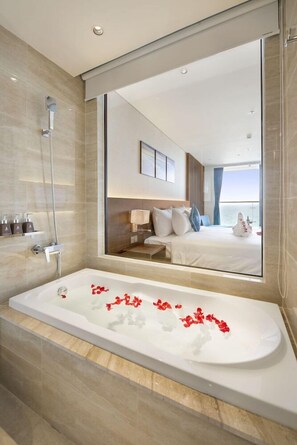 A modern bathroom with a large bathtub, a shower, and a large mirror reflecting a bedroom with a bed and a view of the sea. - Canvas Hotel (Da Nang)