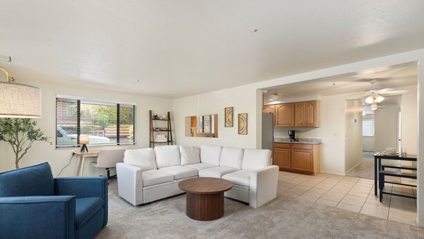A spacious living room with a white sectional sofa, a round coffee table, and a blue armchair. A kitchen is visible in the background with wooden cabinets and a dining table. - Desert Dream Escape – Spacious 2BR Getaway (Sedona)