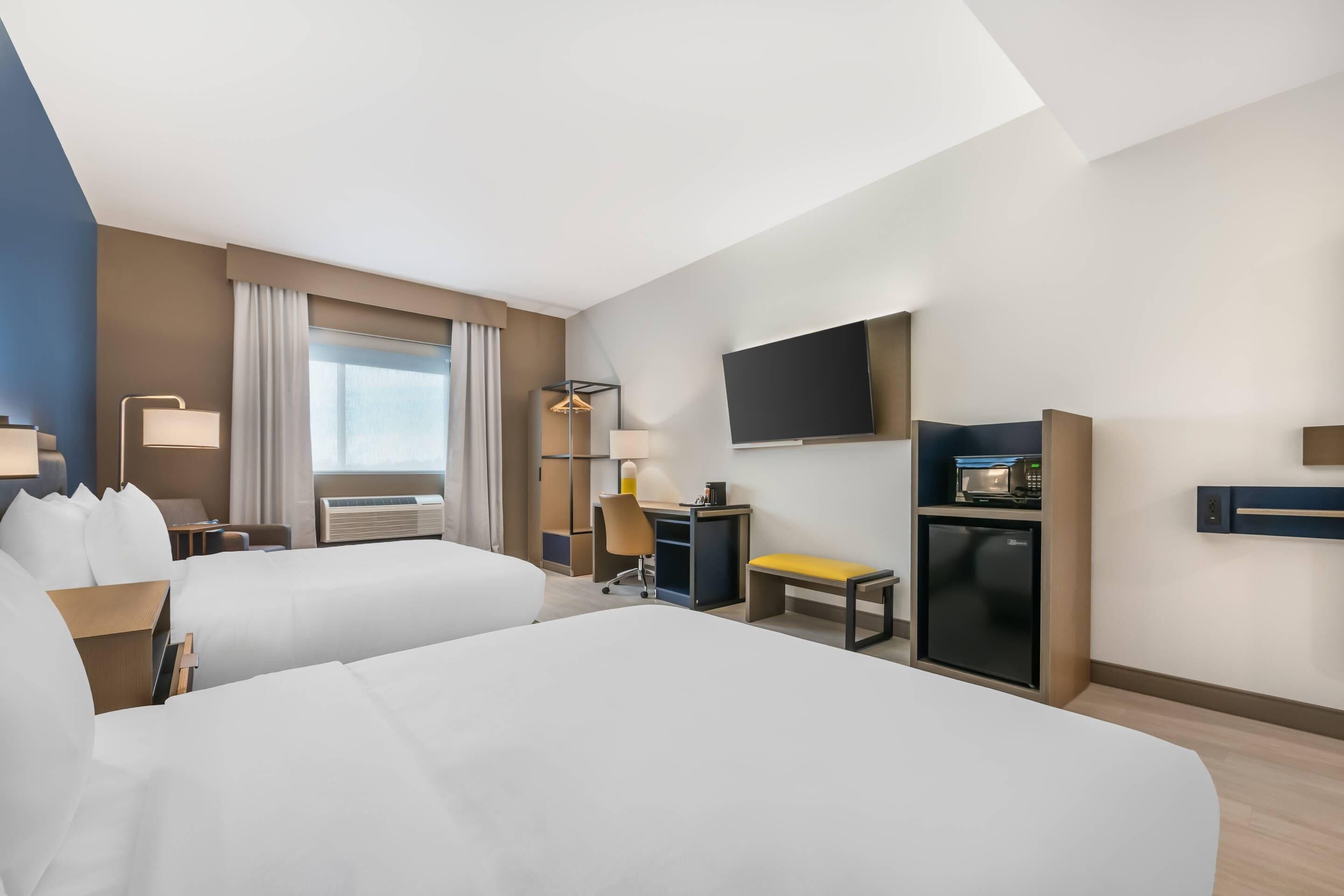 A hotel room with two beds, a desk, a chair, a TV, a mini-fridge, and a window with curtains.