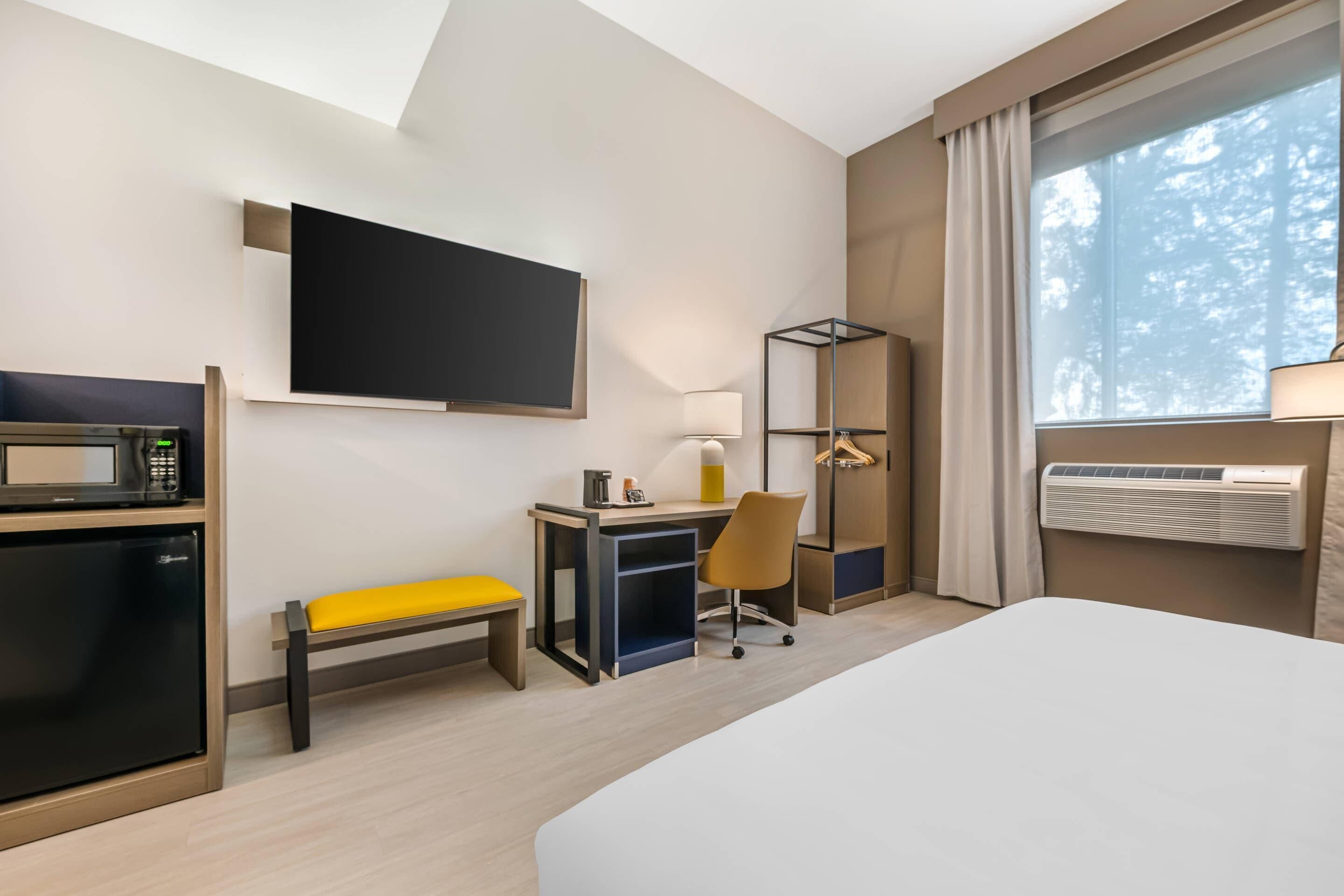 A hotel room with a bed, a desk with a chair, a TV, a microwave, a bench, a lamp, and an air conditioning unit.