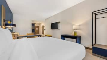 A modern hotel room with a large bed, a desk, a chair, a TV, and a bedside table with a lamp.