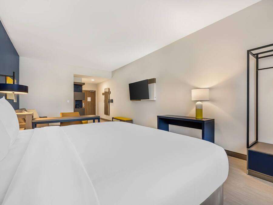 A modern hotel room with a large bed, a desk, a chair, a TV, and a bedside table with a lamp.