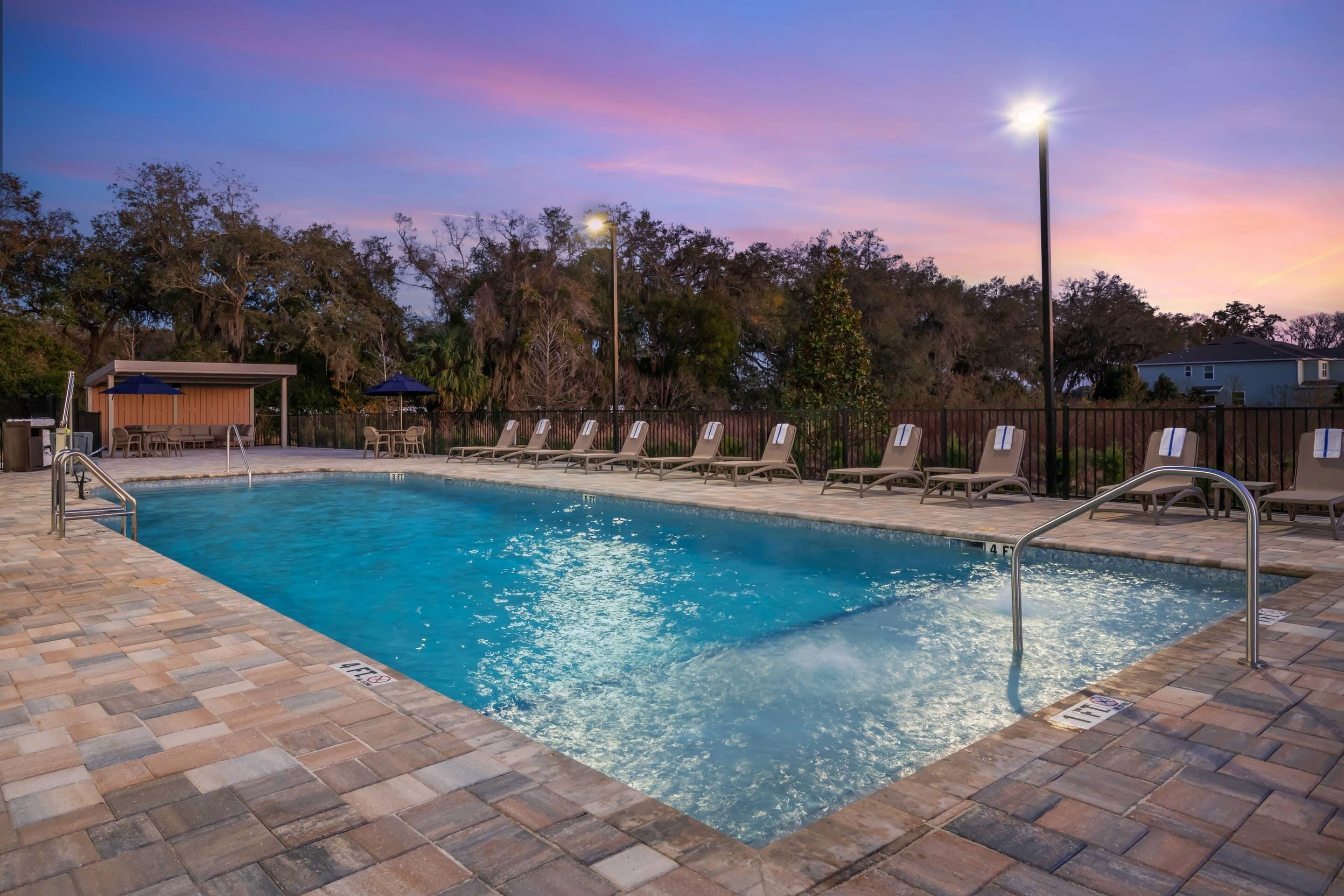 A rectangular swimming pool with a tiled deck, surrounded by lounge chairs and umbrellas, set against a backdrop of trees and a house.