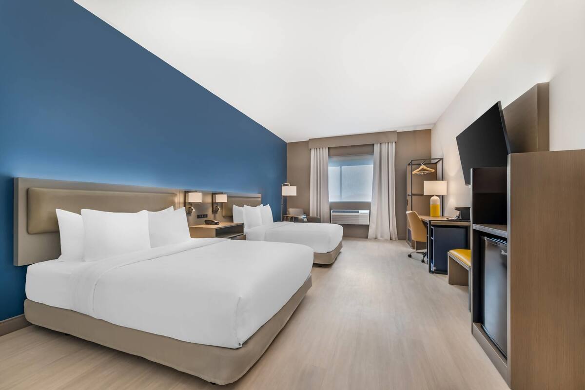 Ett hotellrum med två sängar, ett skrivbord, en stol, en TV och ett fönster med gardiner.