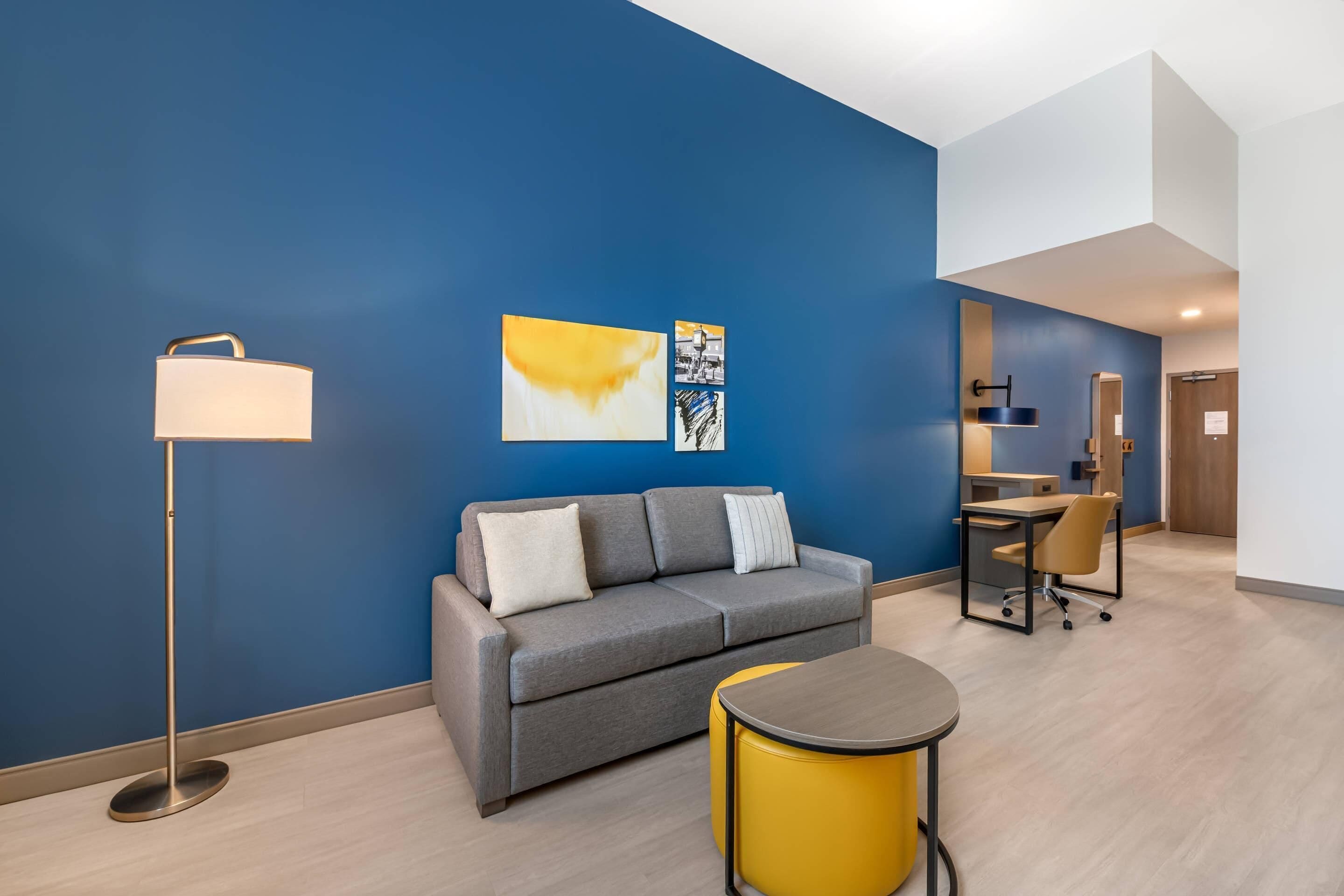 A modern hotel room with a grey sofa, a yellow ottoman, a floor lamp, and artwork on the wall.