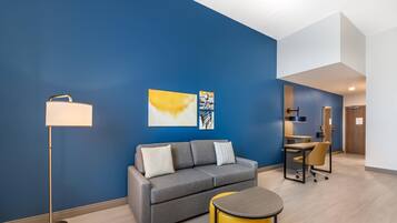 A modern hotel room with a grey sofa, a yellow ottoman, a floor lamp, and artwork on the wall.