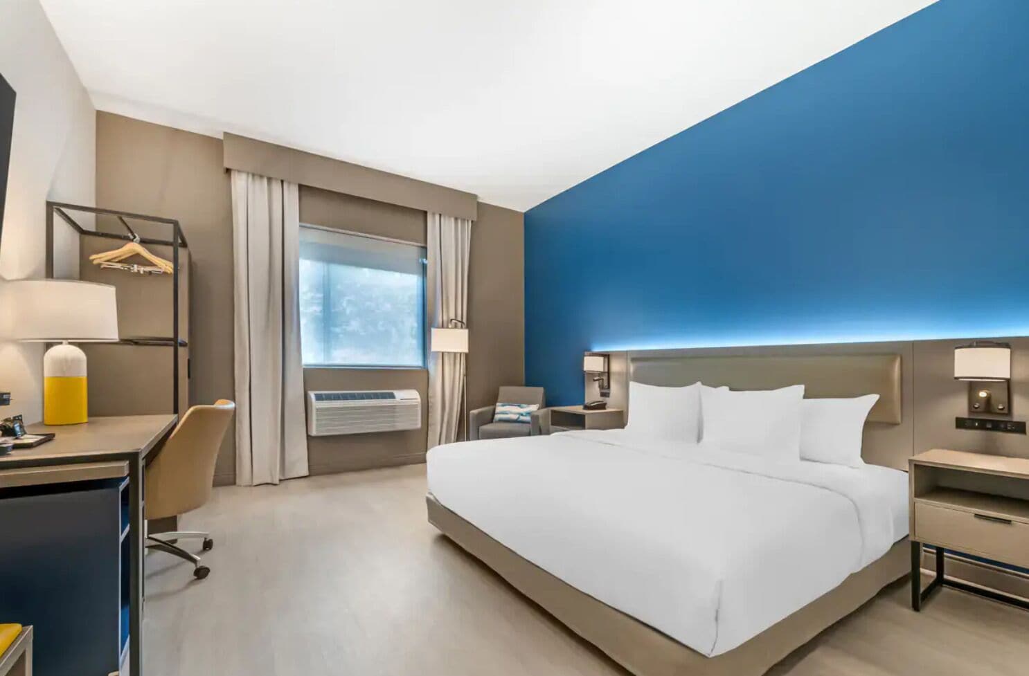 Habitación de hotel con una cama grande, un escritorio, una silla, una mesita de noche, una lámpara y un equipo de aire acondicionado.