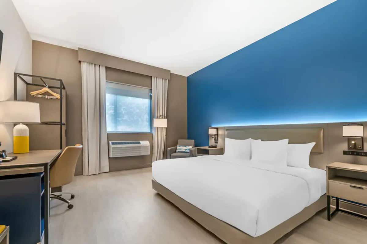 Ett hotellrum med en stor säng, ett skrivbord, en stol, ett nattduksbord, en lampa och en luftkonditioneringsanläggning.