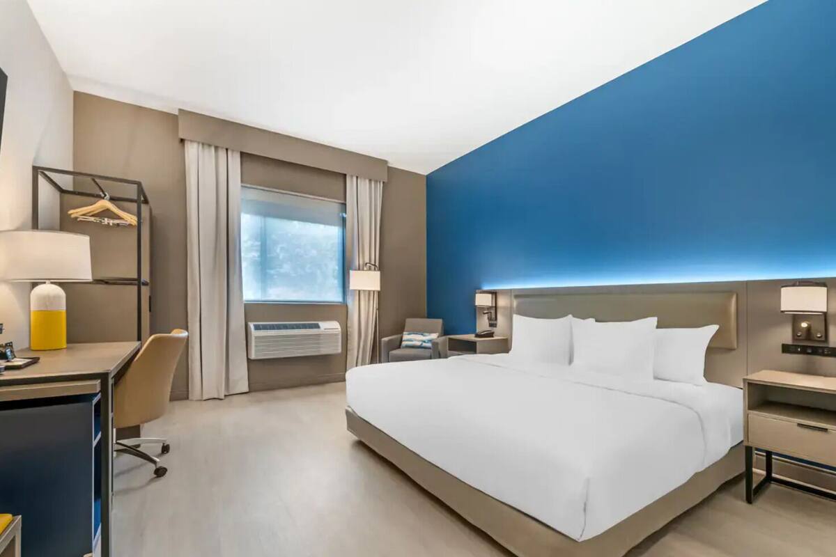 Ett hotellrum med en stor säng, ett skrivbord, en stol, ett nattduksbord, en lampa och en luftkonditioneringsanläggning.