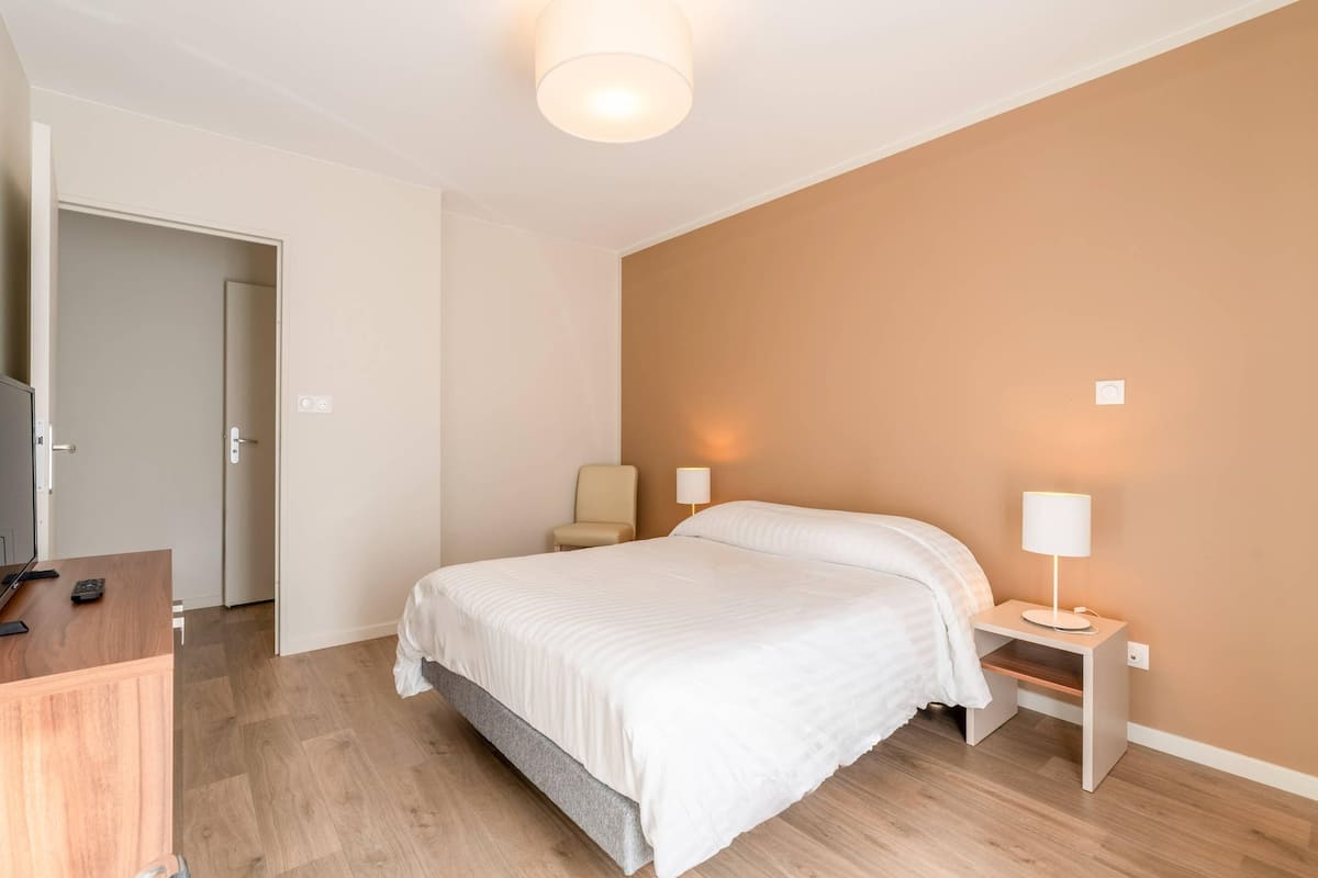 Un dormitorio con cama, mesitas de noche, una silla y un escritorio con lámpara.