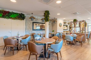 A modern restaurant with round tables and blue chairs, a green wall decoration, and a wooden counter. - Quality Aparthotel Grenoble Sud (Echirolles)