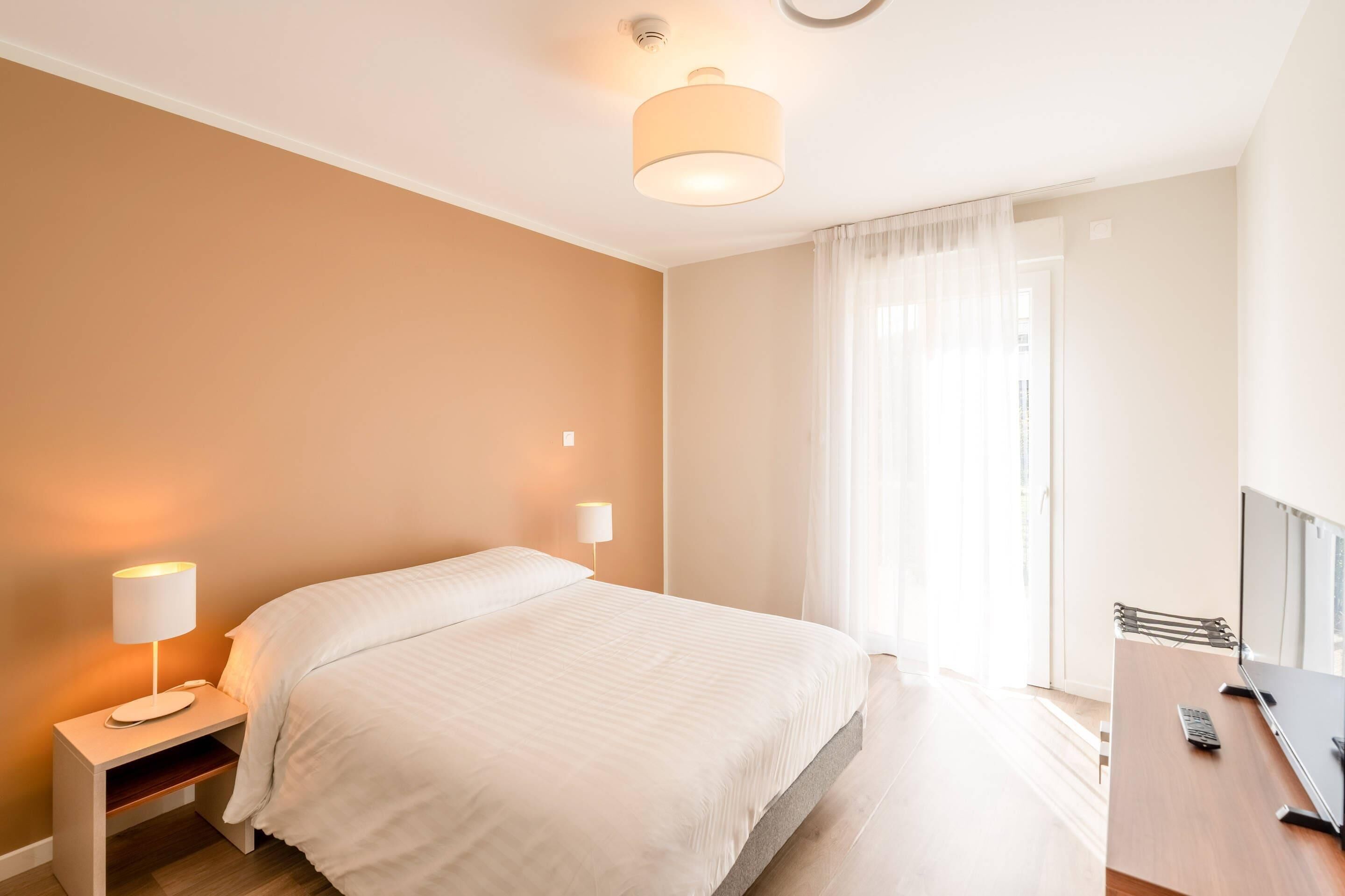 A hotel room with a bed, bedside table, lamp, and a desk with a chair.