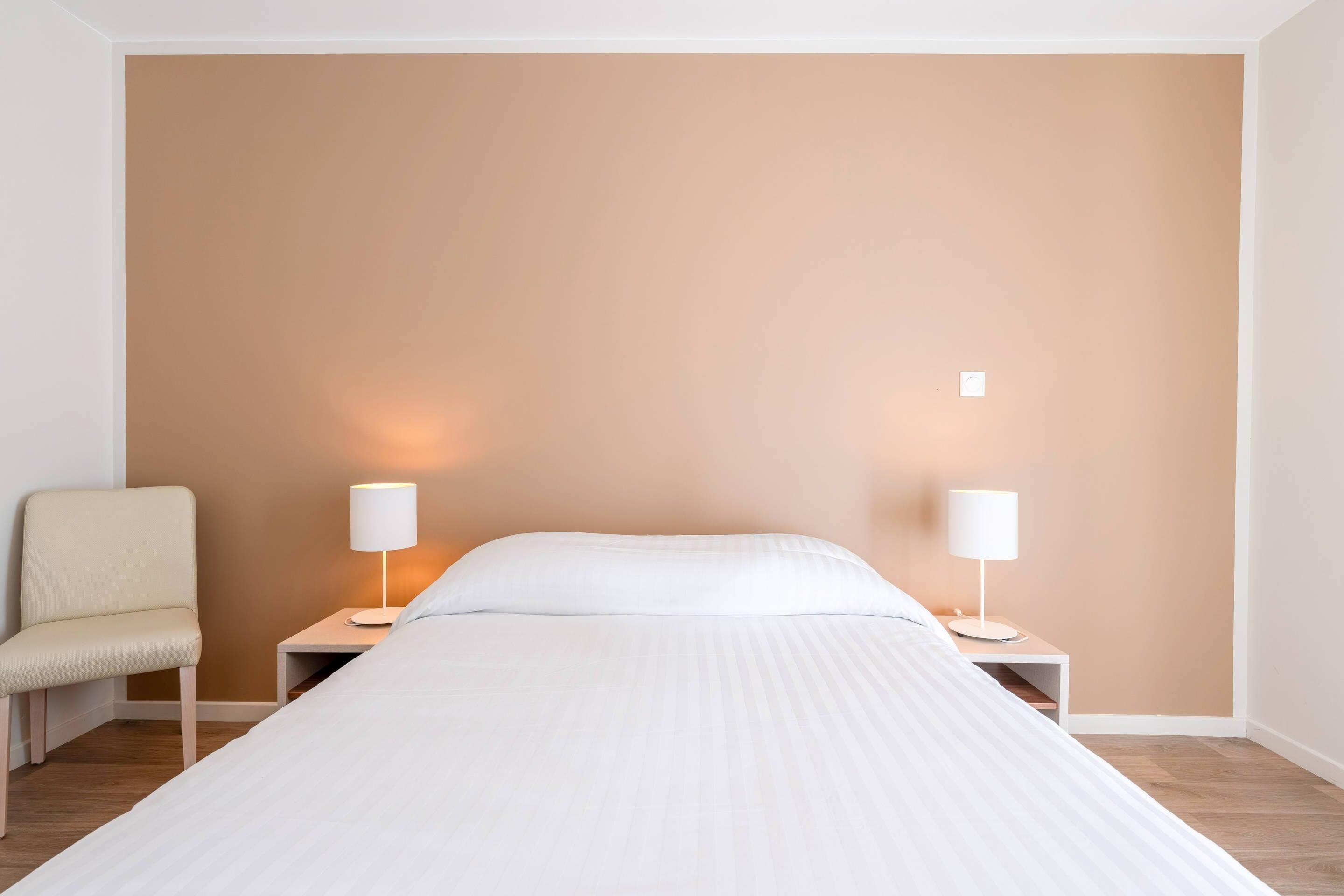 A hotel room with a bed, two bedside tables with lamps, a chair, and a plain wall.
