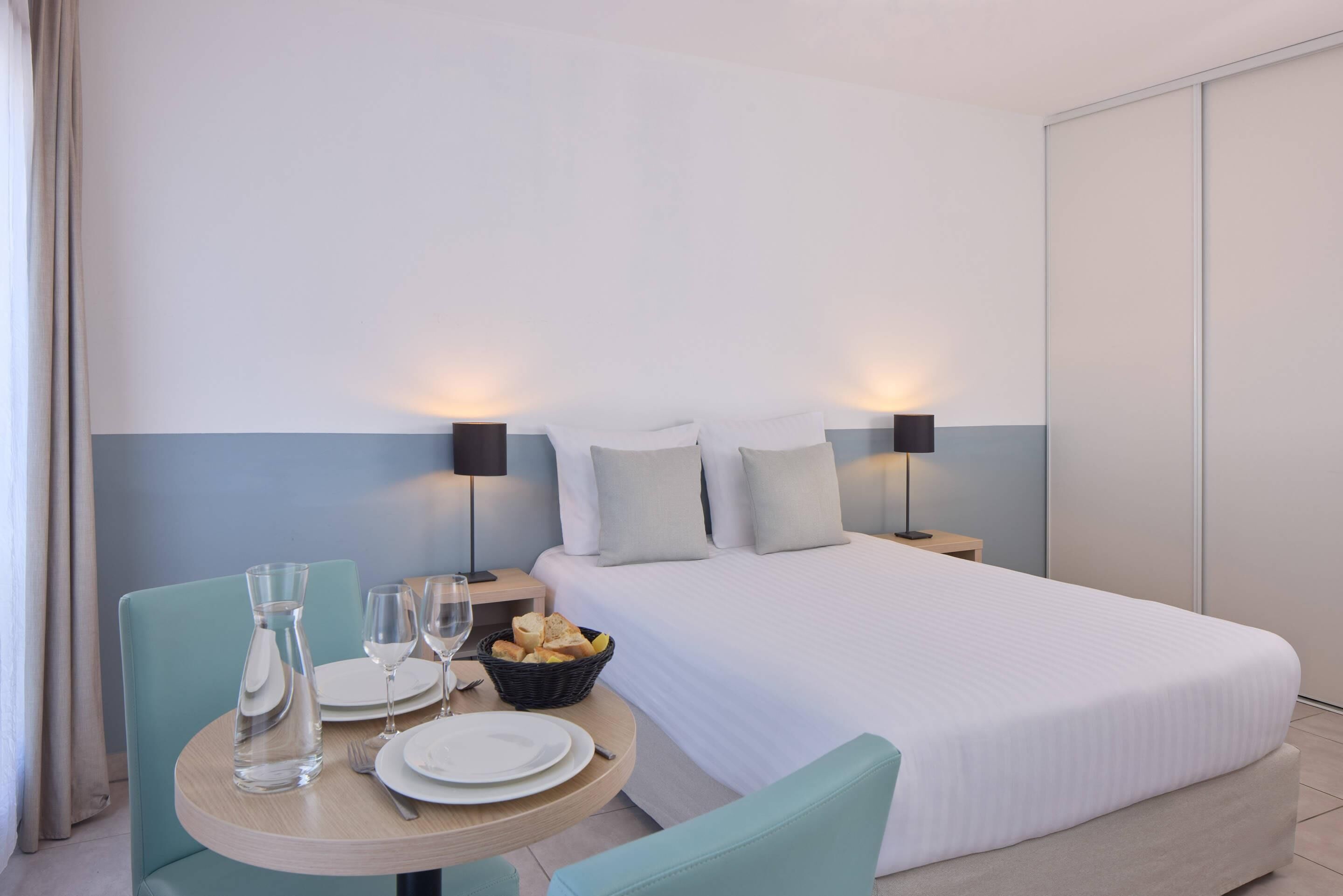 A hotel room with a bed, a round dining table with chairs, a basket of bread, and two bedside lamps.