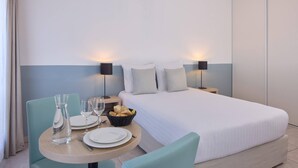 Une chambre d’hôtel avec un lit, une table de salle à manger ronde entourée de chaises, un panier de pain et deux lampes de chevet.