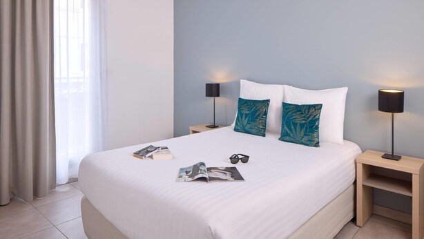 Un lit bien fait, avec du linge de lit blanc, deux coussins décoratifs, une paire de lunettes et un magazine posés sur le lit. Il y a deux tables de chevet avec des lampes, une de chaque côté du lit.