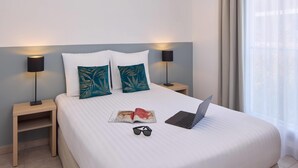 Un lit bien fait, avec un ordinateur portable, des lunettes de soleil et un magazine, dans une chambre d’hôtel.