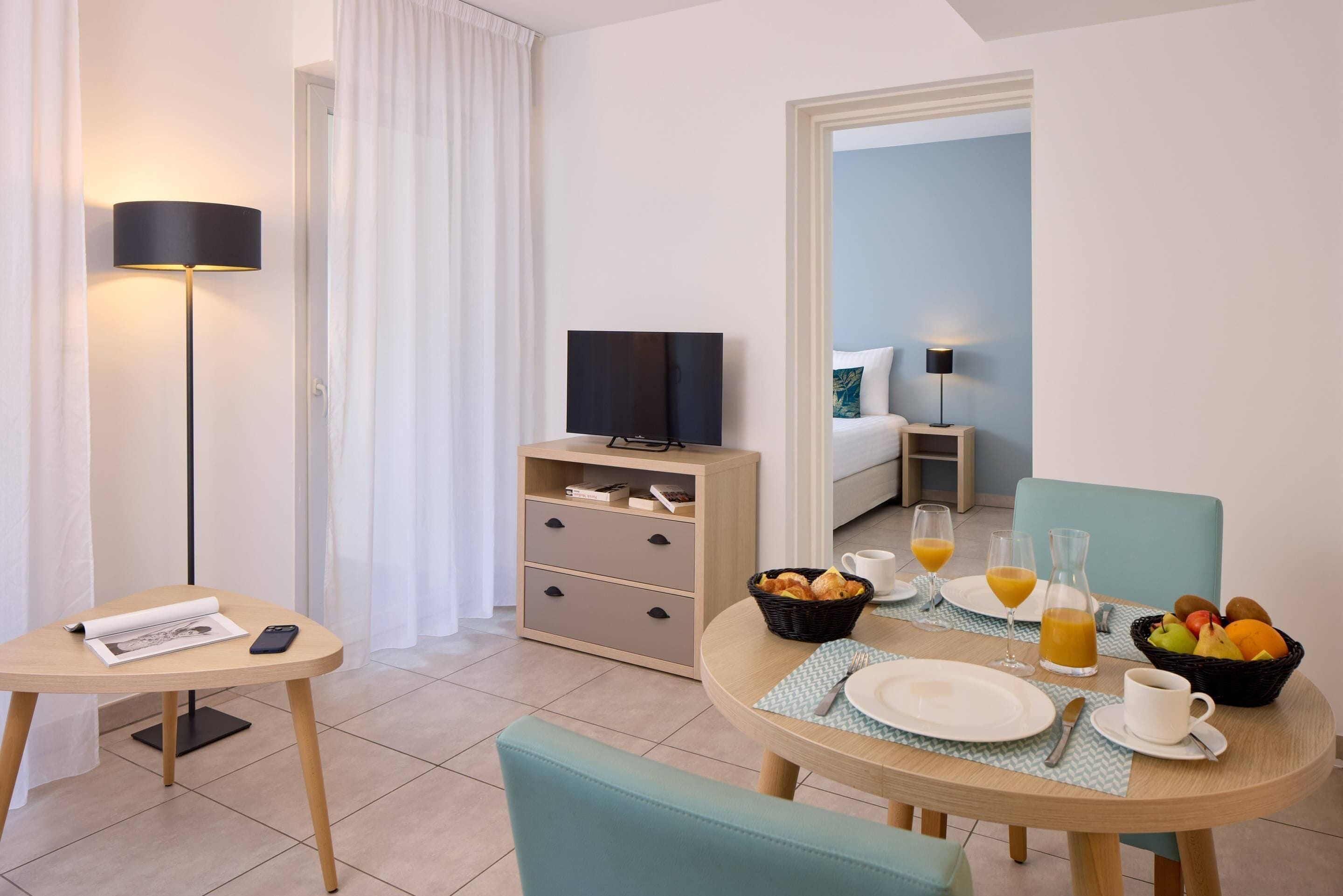 A hotel room with a dining table set for breakfast, a TV, a bedside table, and a bed in the background.