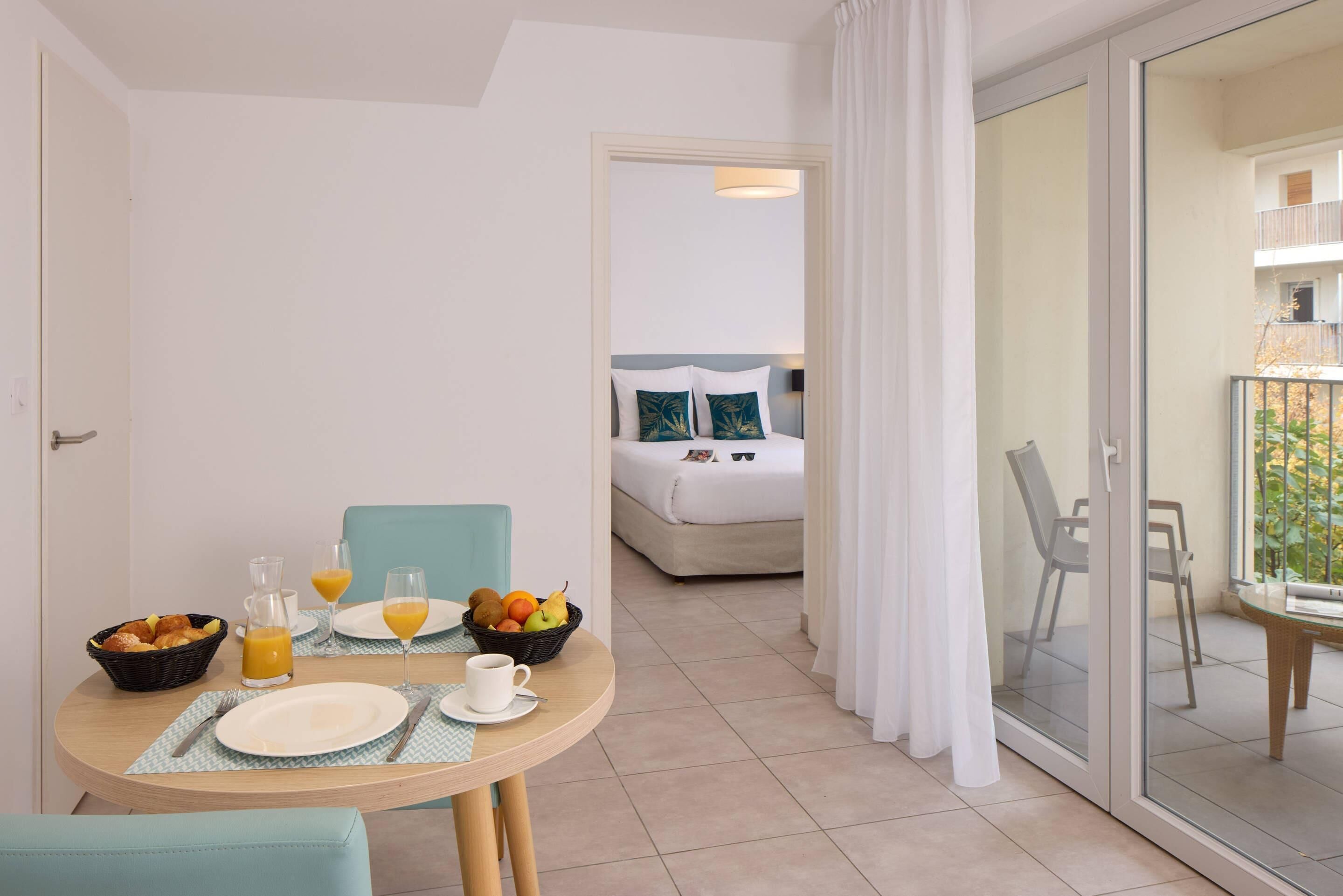 A hotel room with a breakfast setup including a table with plates, glasses, and a fruit bowl. There is a bed with pillows and a view of the balcony.
