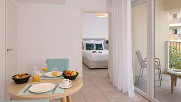 A hotel room with a breakfast setup including a table with plates, glasses, and a fruit bowl. There is a bed with pillows and a view of the balcony.