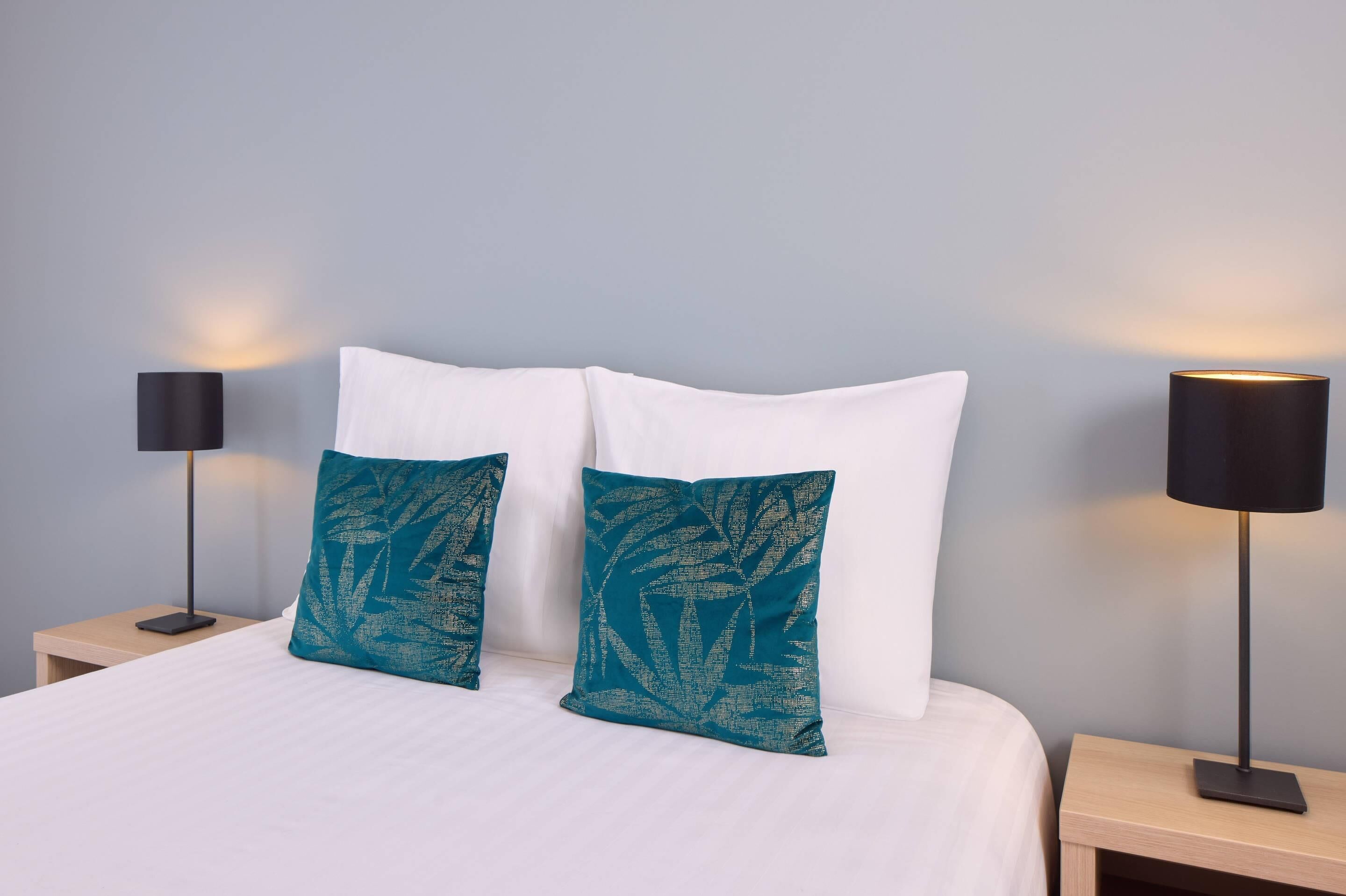 A neatly made bed with white bedding and two teal pillows, flanked by two black bedside tables with lamps.