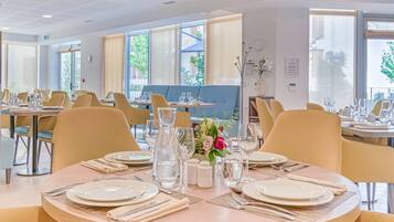 A dining area with round tables, beige chairs, and a view of the outdoors through large windows.