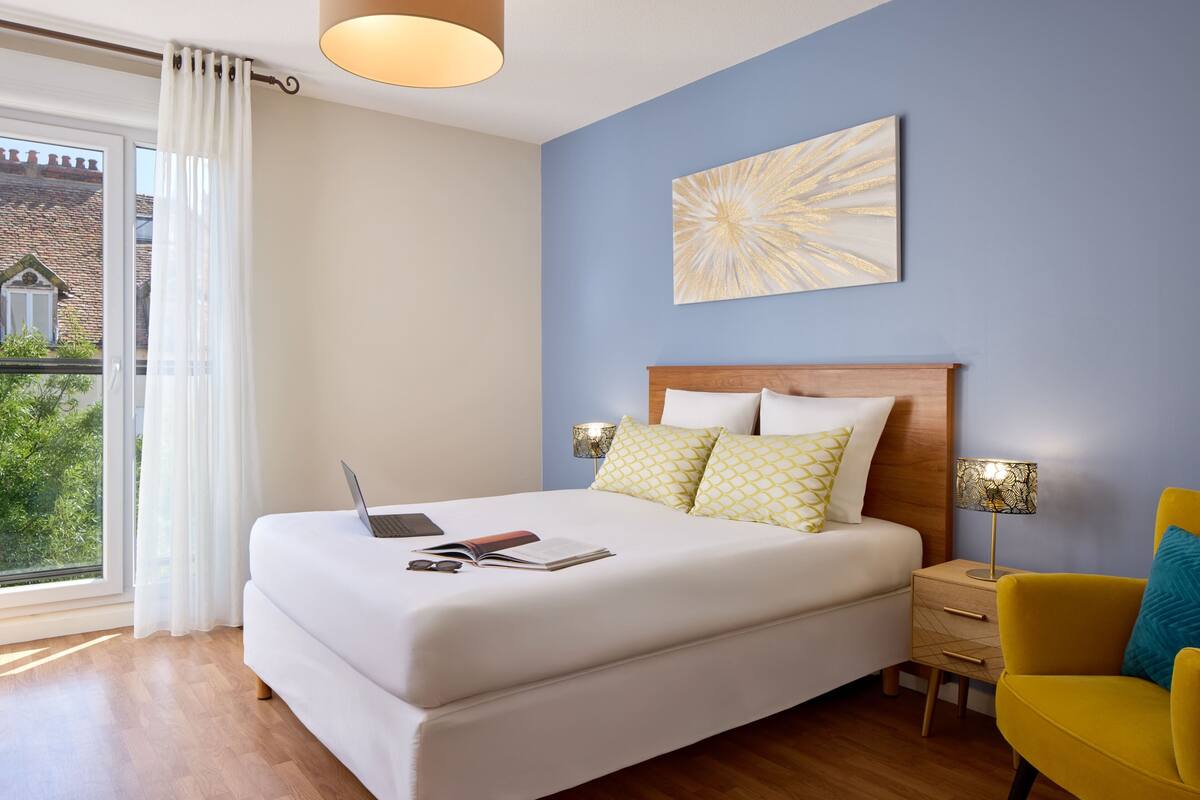 A bedroom with a bed, a laptop, a bedside table, a lamp, and a view of greenery outside.