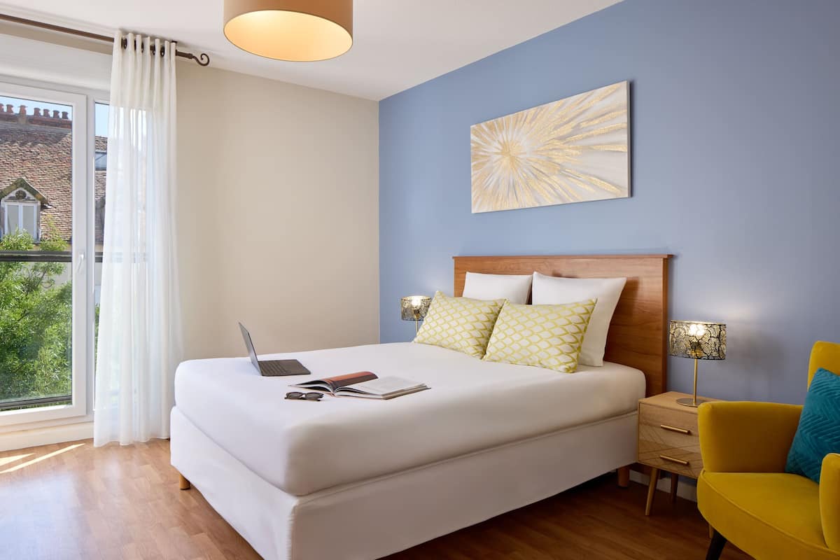 Une chambre à coucher avec un lit, un ordinateur portable, une table de chevet, une lampe et une vue sur la verdure.