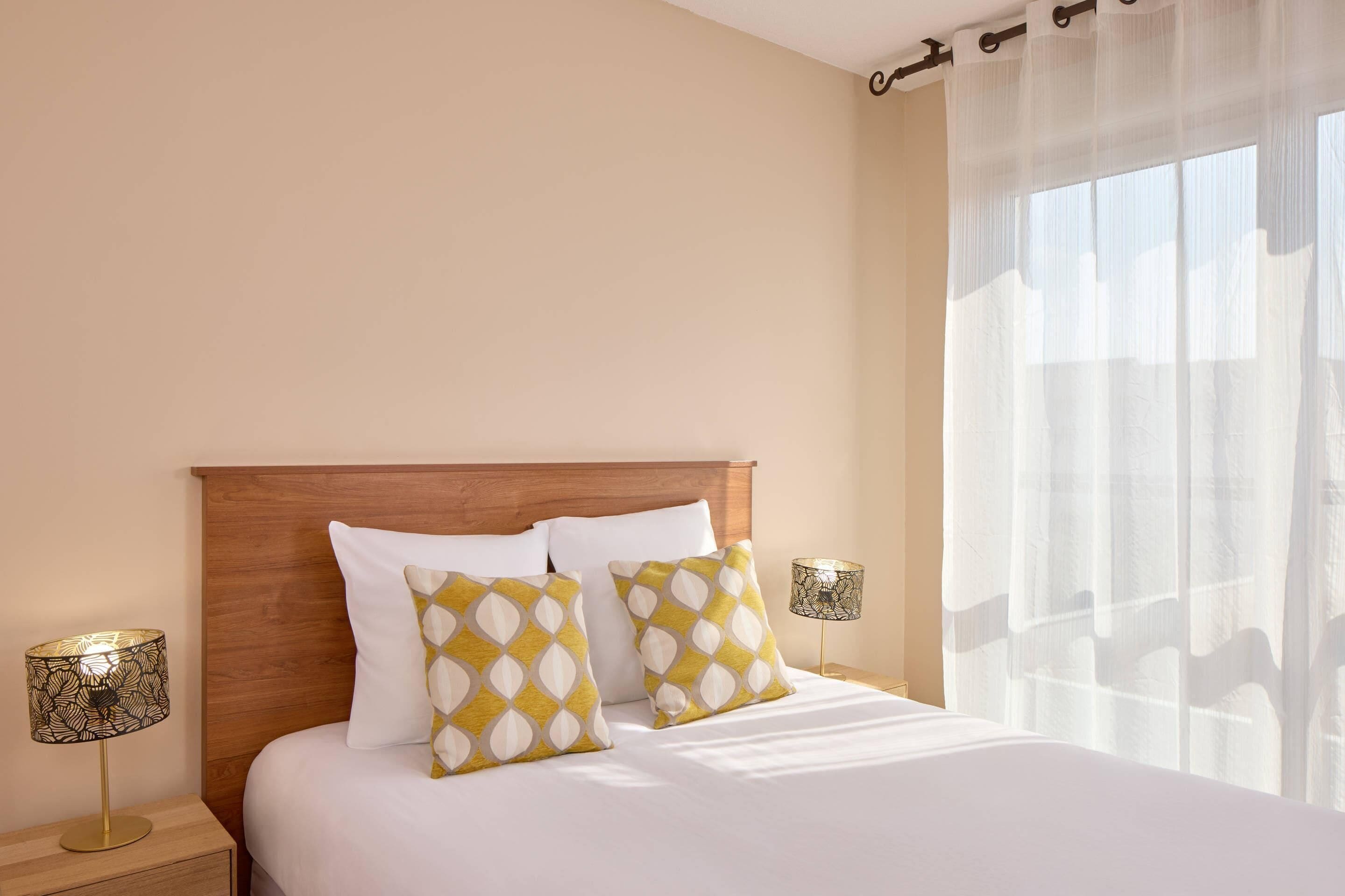 A neatly made bed with white bedding and two patterned pillows, a wooden headboard, and two bedside lamps with lampshades.