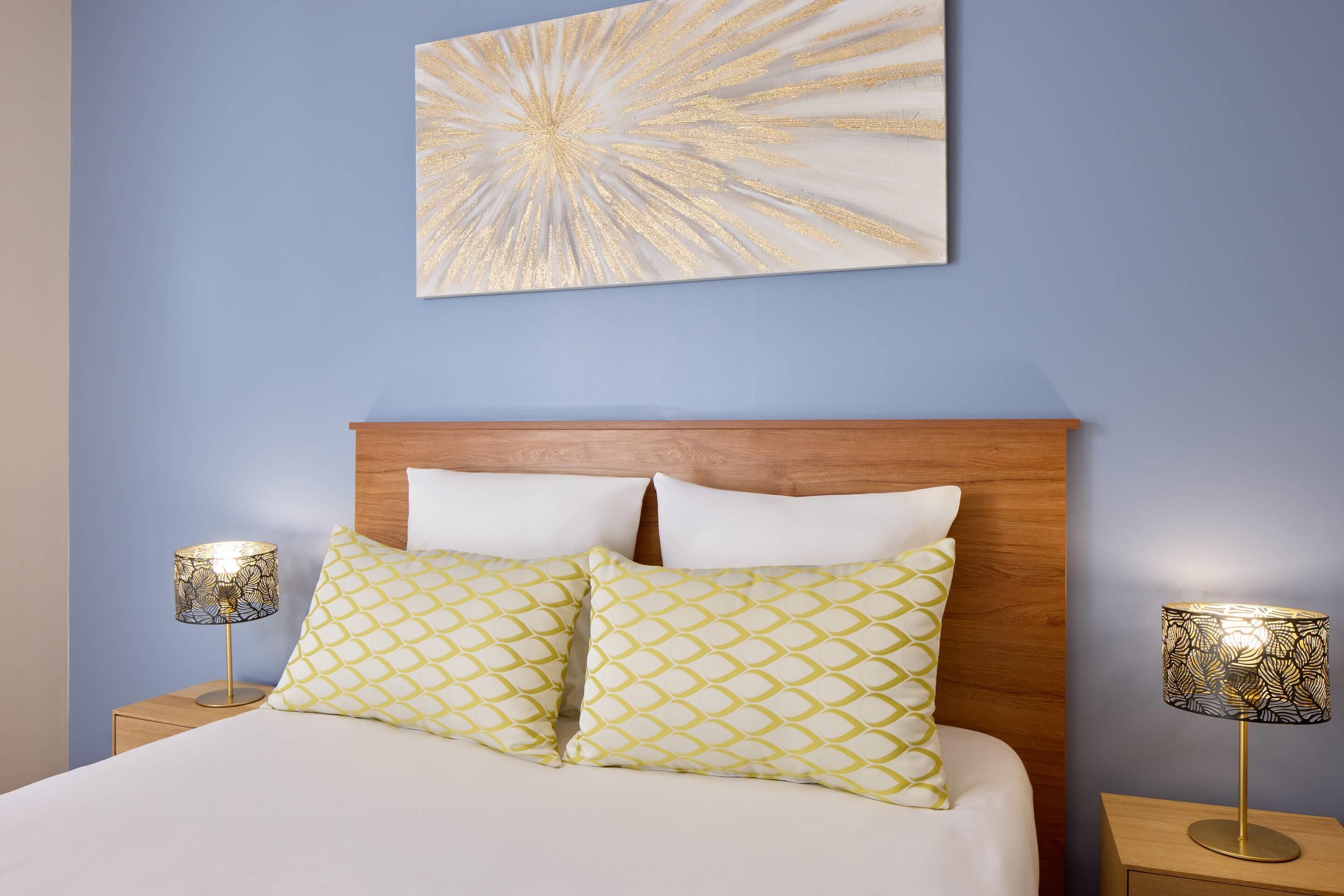 A neatly made bed with two patterned pillows, a wooden headboard, and two bedside lamps with patterned shades.