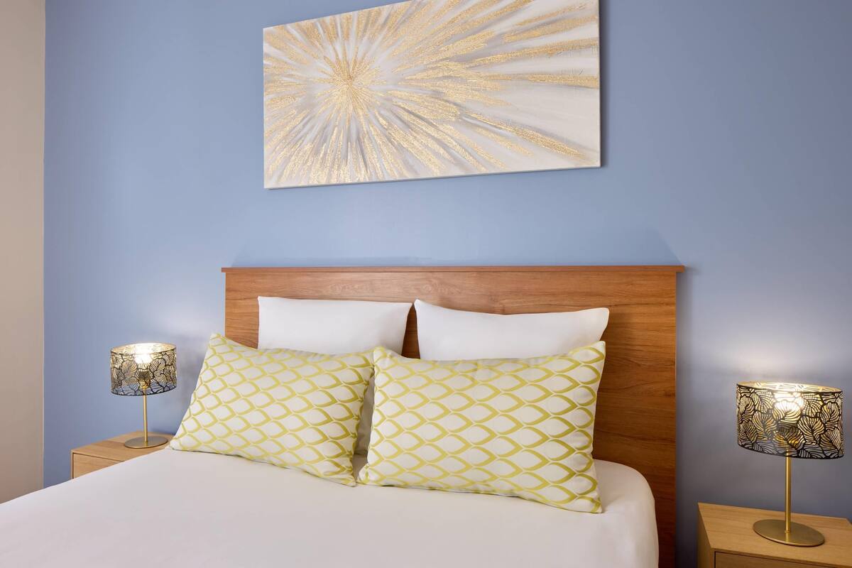 A neatly made bed with two patterned pillows, a wooden headboard, and two bedside lamps with patterned shades.