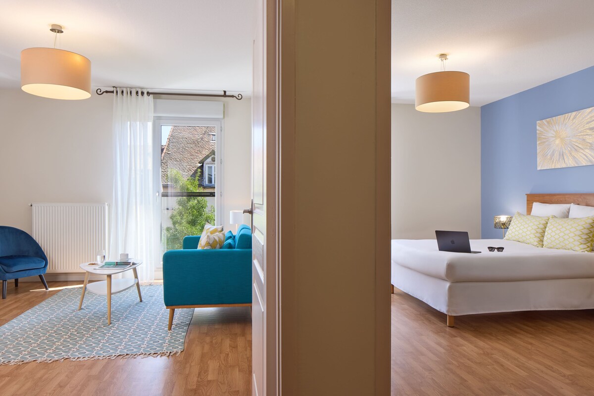 Habitación de hotel moderna con una cama, un sillón azul, una mesita y vistas al exterior.