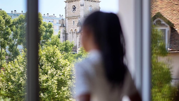 A person looking out of a window at a historic tower with a clock, surrounded by greenery. - Quality Aparthotel Chalon-sur-Saône Gare (Chalon-sur-Saone)