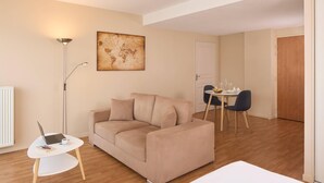 A modern living room with a beige sofa, a small white table with a laptop, and a dining area with a round table and chairs. - Quality Aparthotel Chalon-sur-Saône Gare (Chalon-sur-Saone)