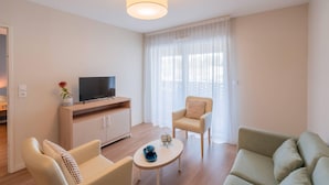 A modern living room with a sofa, armchair, and a small coffee table. A TV is on a wooden cabinet, and a window with sheer curtains is visible. - Quality Aparthotel Toulouse Ponts Jumeaux (Toulouse)