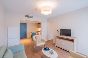 Un salon moderne avec un canapé, une table basse, un téléviseur posé sur un meuble en bois, et un coin repas à l’arrière-plan.