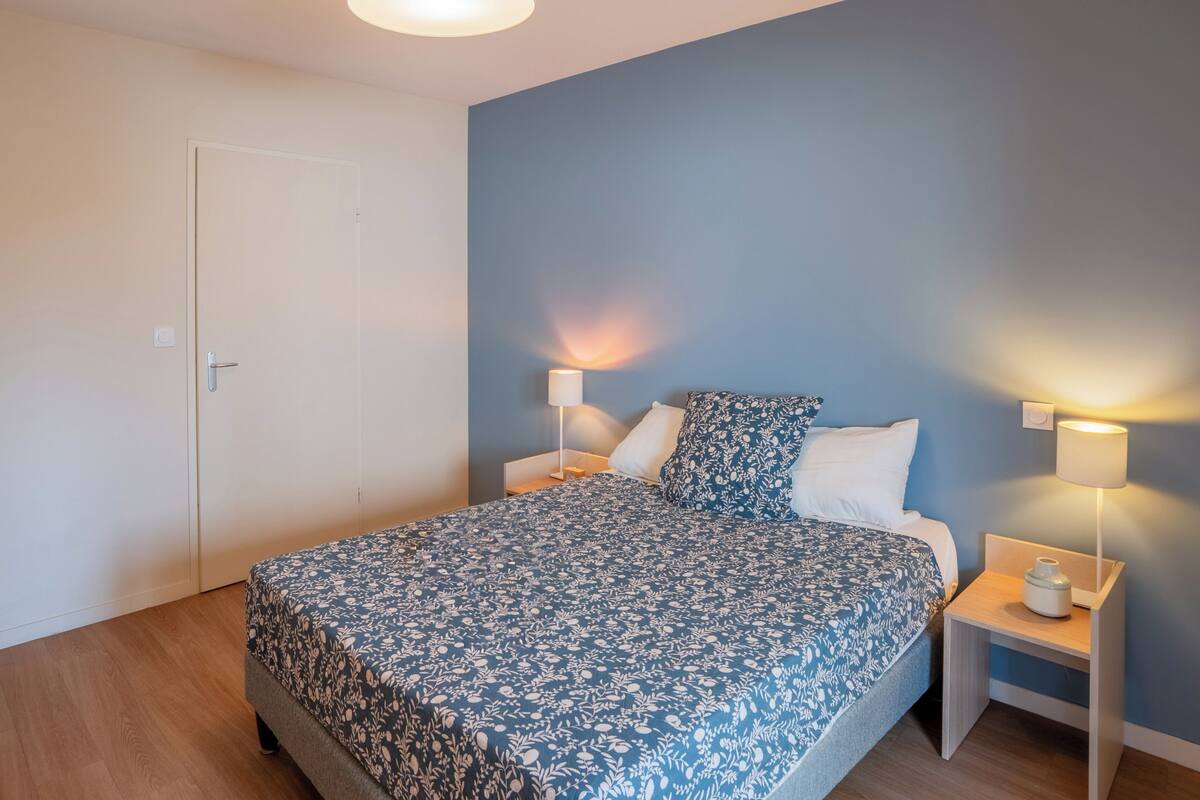 Une chambre à coucher comprenant un lit, deux tables de chevet, une commode, une porte et un luminaire fixé au mur.