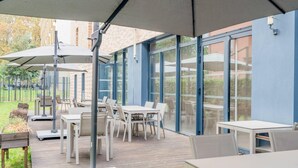 Ein Außenbereich mit einer Terrasse, Tischen, Stühlen und großen Sonnenschirmen, mit Blick auf die Umgebung durch Glastüren.