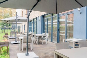 An outdoor patio area with tables and chairs, large umbrellas, and a view of the surrounding area through glass doors. - Quality Aparthotel Toulouse Ponts Jumeaux (Toulouse)