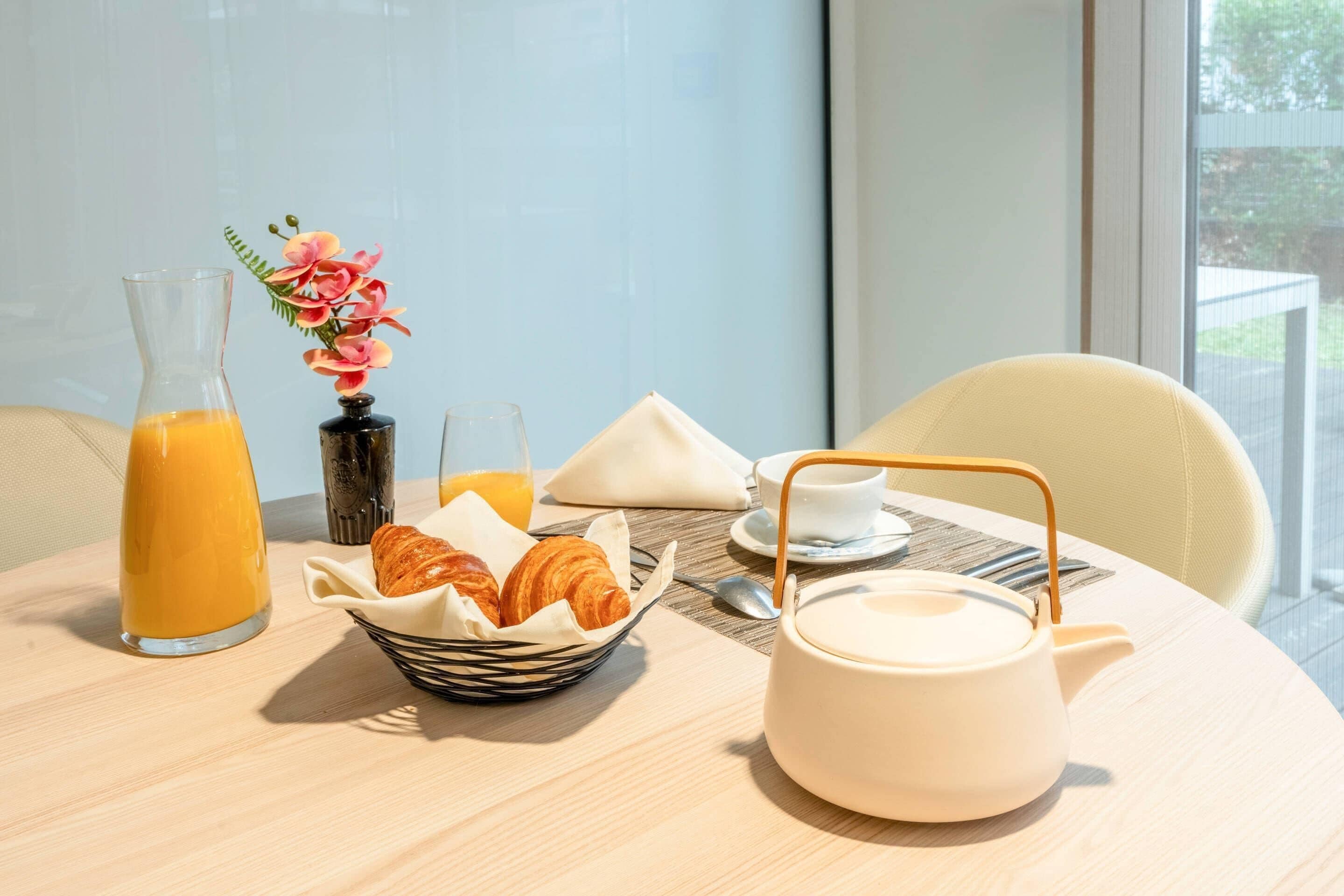 Une table en bois dressée pour le petit-déjeuner, avec des croissants, du jus d’orange et une théière.
