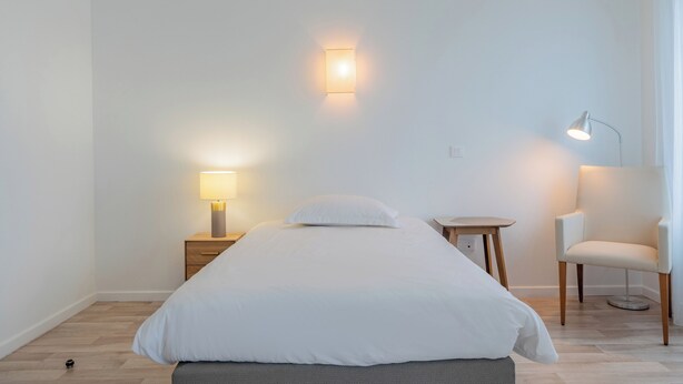A minimalist bedroom with a bed, bedside tables, a chair, and wall-mounted lighting. - Quality Aparthotel Mulhouse (Mulhouse)