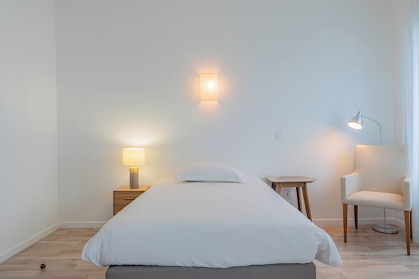A minimalist bedroom with a bed, bedside tables, a chair, and wall-mounted lighting. - Quality Aparthotel Mulhouse (Mulhouse)