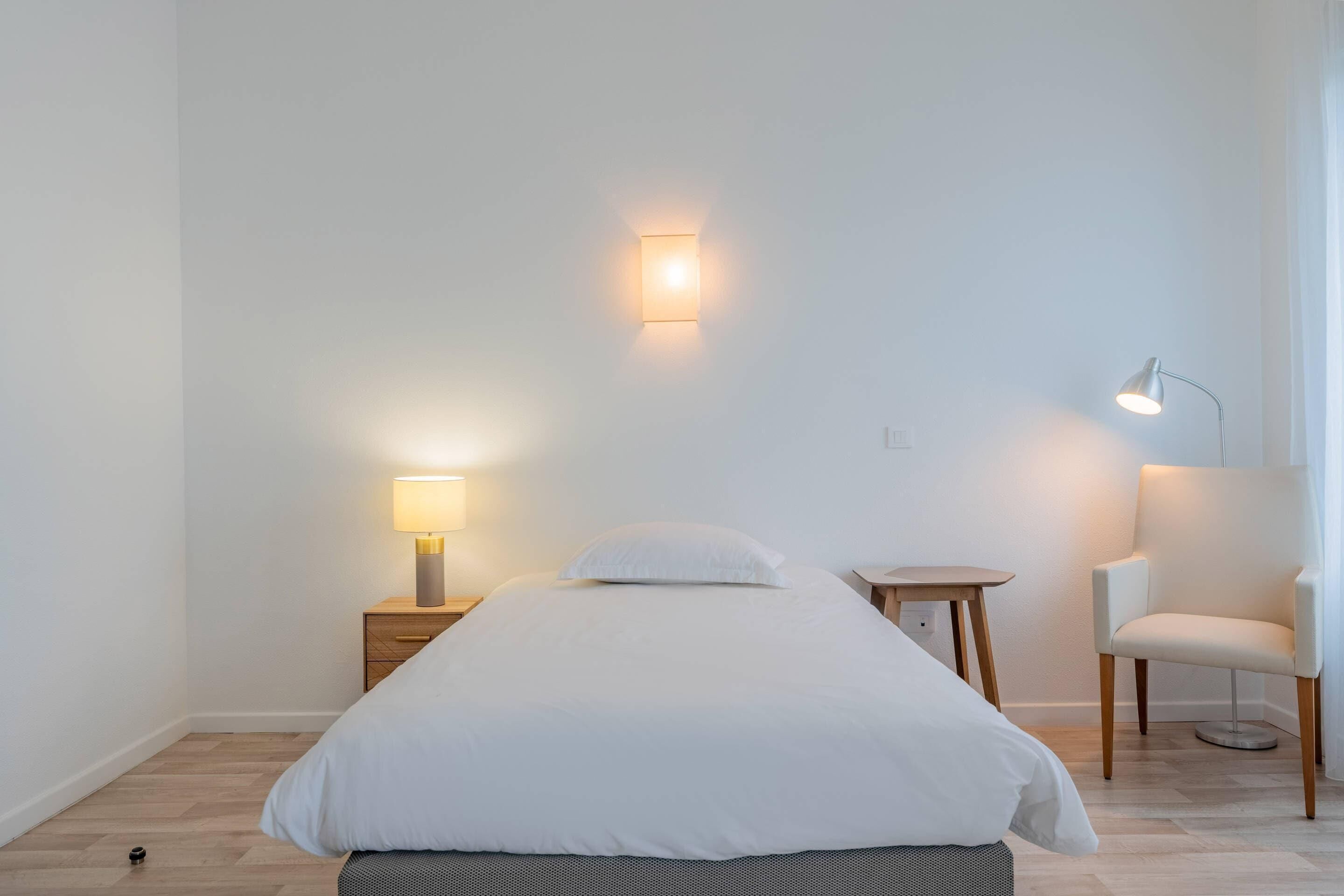 A minimalist bedroom with a bed, bedside table, lamp, chair, and wall-mounted light fixture.